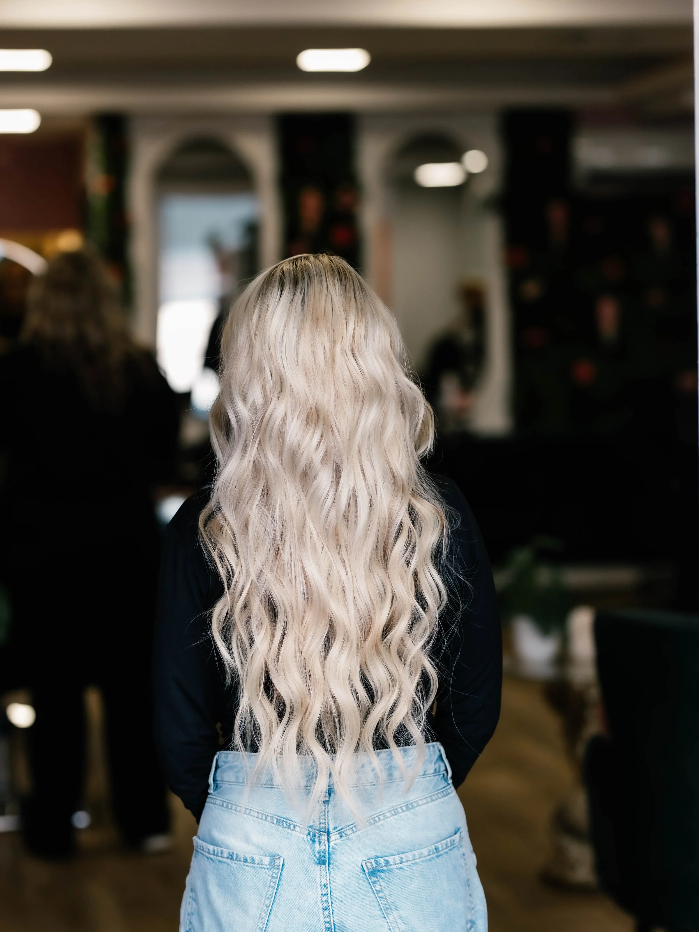 Klant met lang ijsblond golvend haar, van achteren gezien in de salon