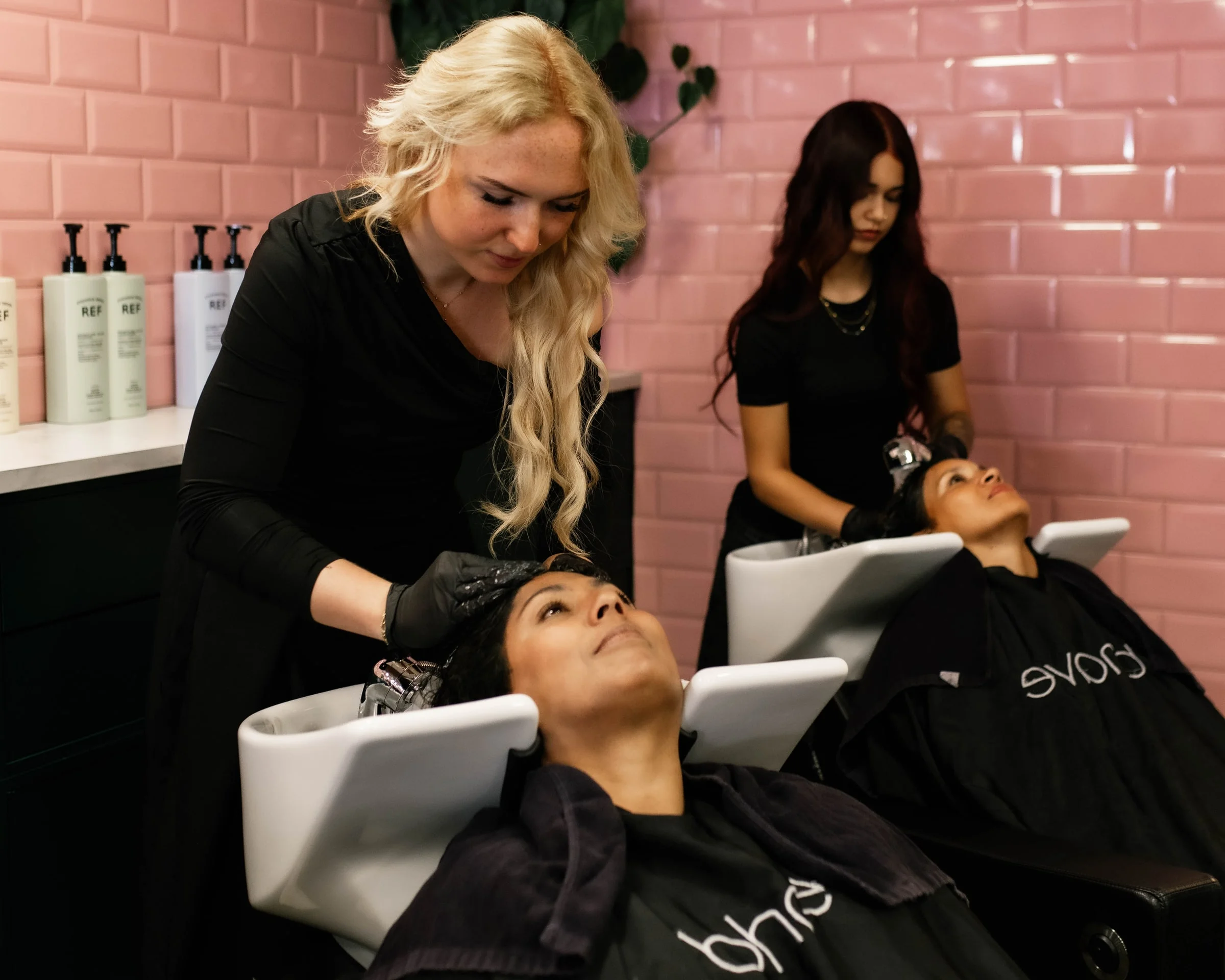 Twee stylisten wassen het haar van klanten bij de wasbakken tegen de roze tegelwand